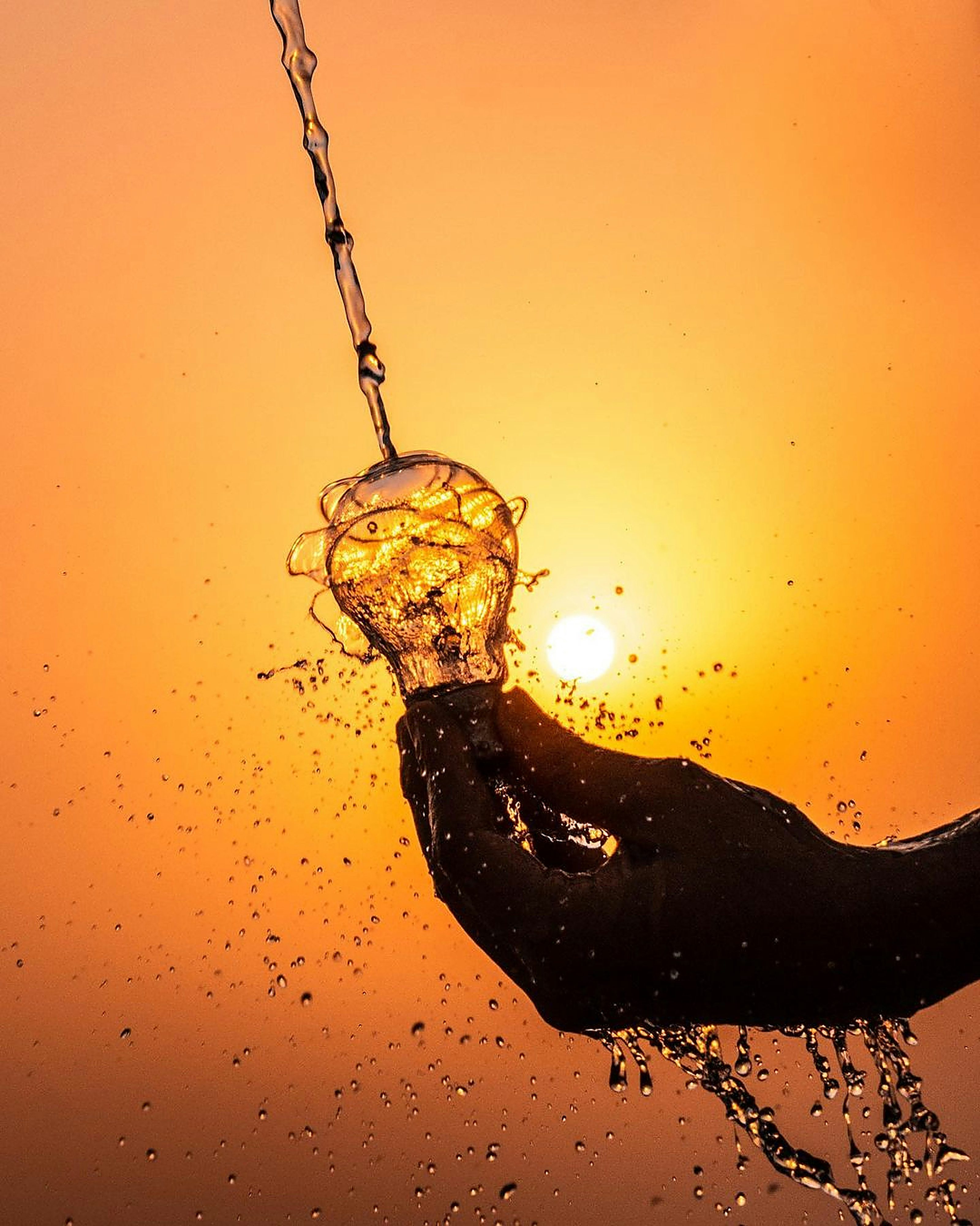 A hand holding a light bulb under pouring water against an orange sunset